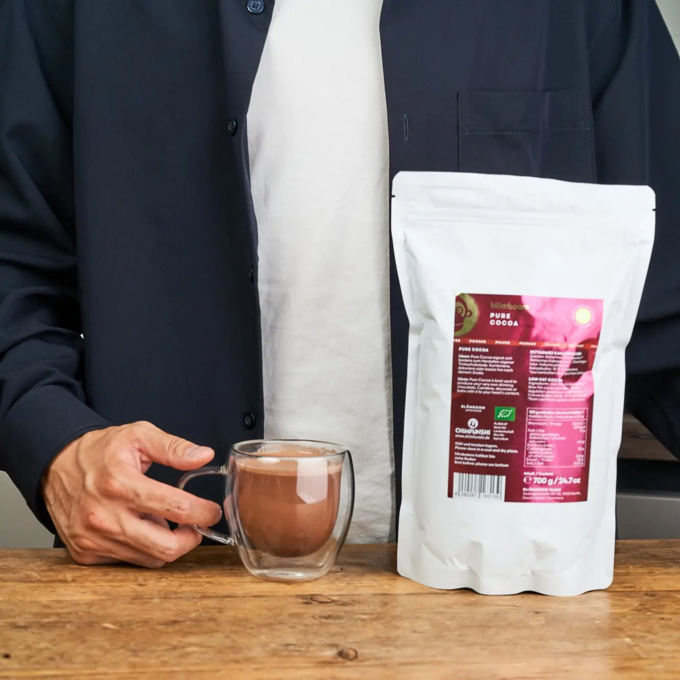 Ekologiczne kakao Blömboom Pure Cocoa 700g – opakowanie i filiżanka z napojem trzymana przez osobę przy stole