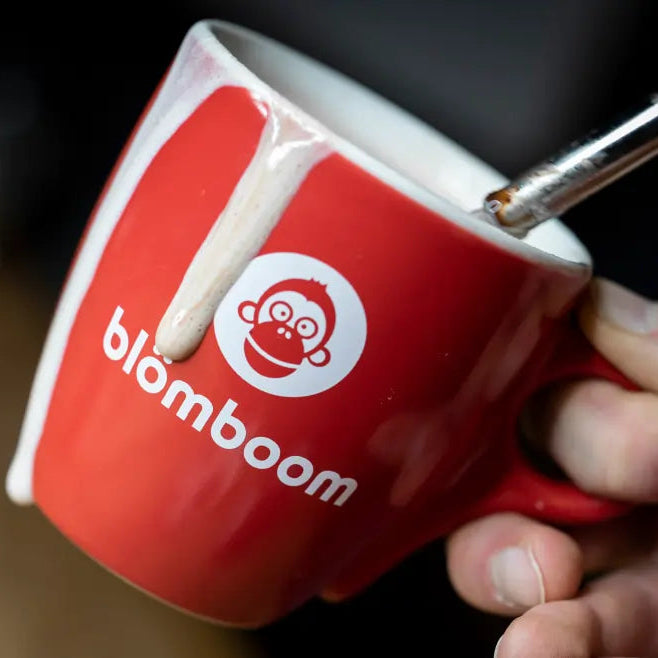 Czerwona filiżanka z logo "blömboom" i grafiką małpy, z której spływa mleczna piana, trzymana w dłoni baristy.