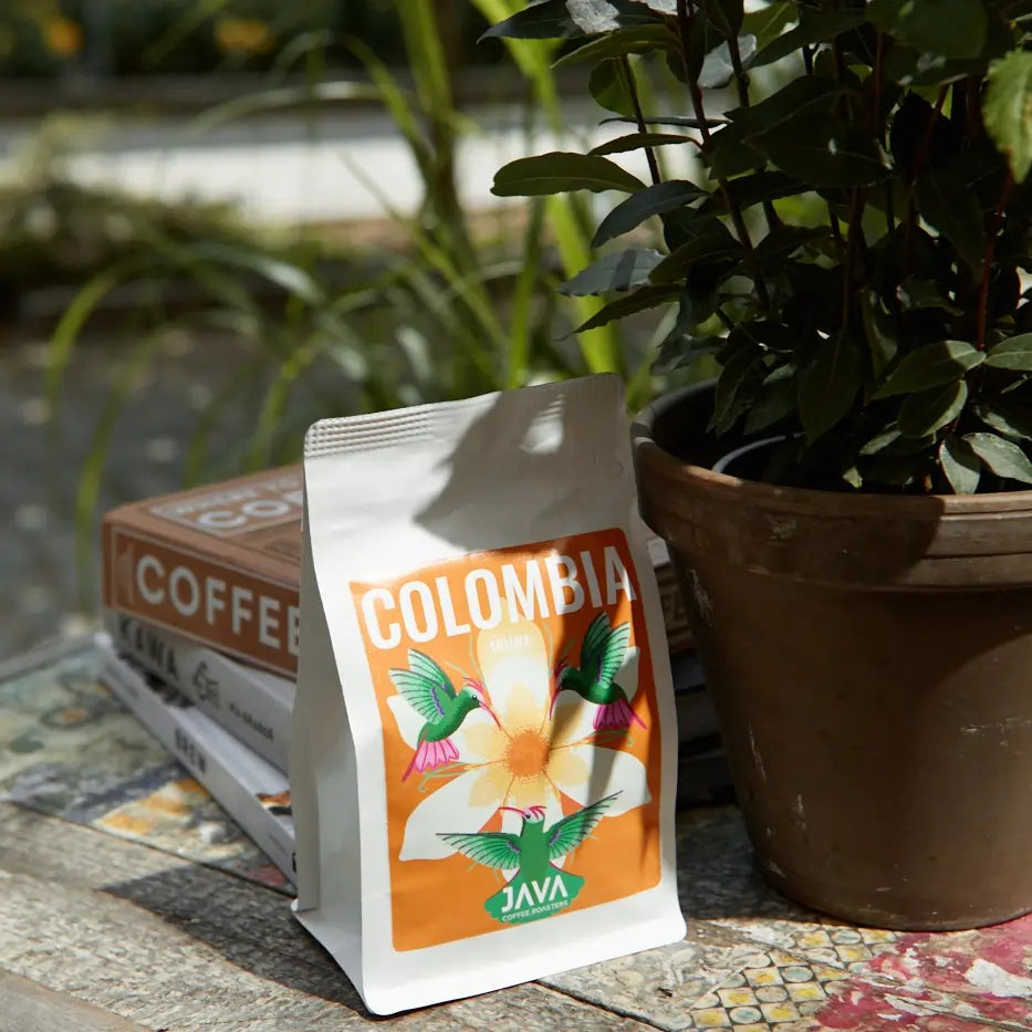 Kawa Colombia Tolima JAVA Coffee Roasters – opakowanie ustawione obok książek o kawie i doniczki z rośliną na rustykalnym stole