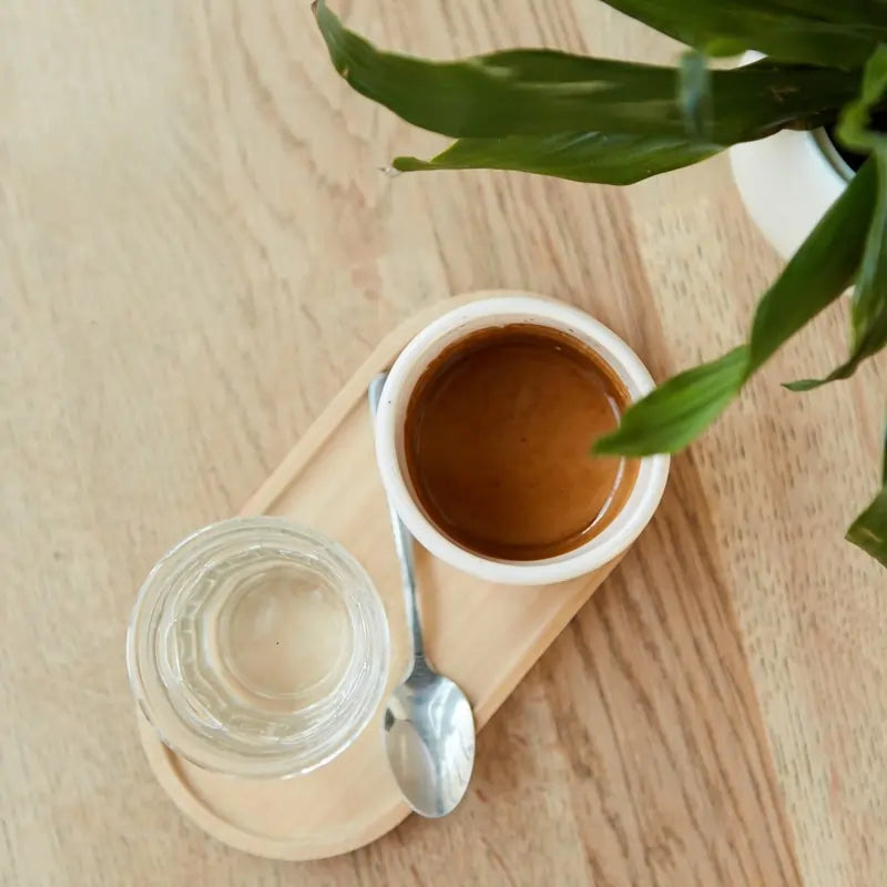 Filiżanka espresso na drewnianej tacy, obok szklanki wody i łyżeczki, z zieloną rośliną w tle.
