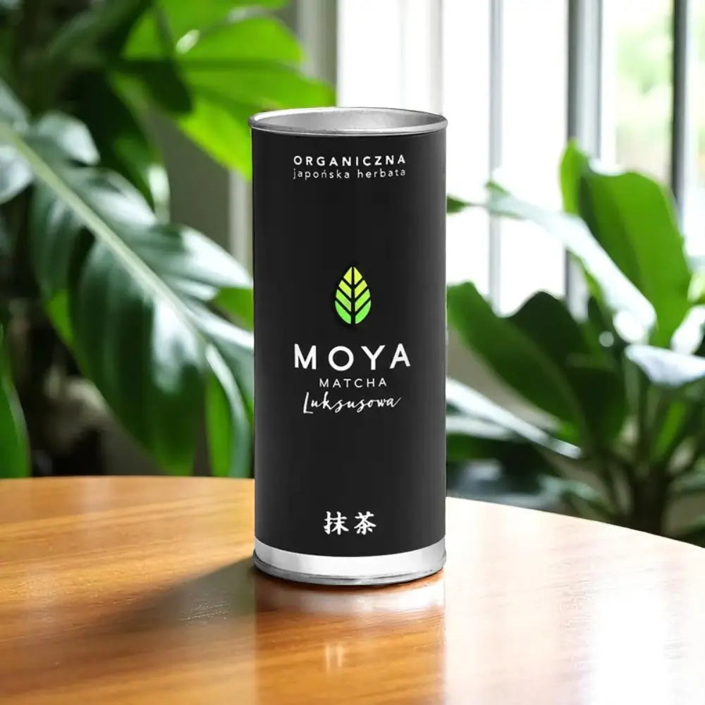 Herbata MOYA Matcha Luksusowa w czarnym opakowaniu na tle zielonych roślin – organiczna japońska matcha premium
