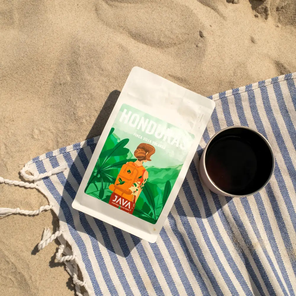 Opakowanie kawy ziarnistej Honduras Finca Villa Yolanda JAVA Coffee Roasters na plaży, obok kubka z kawą