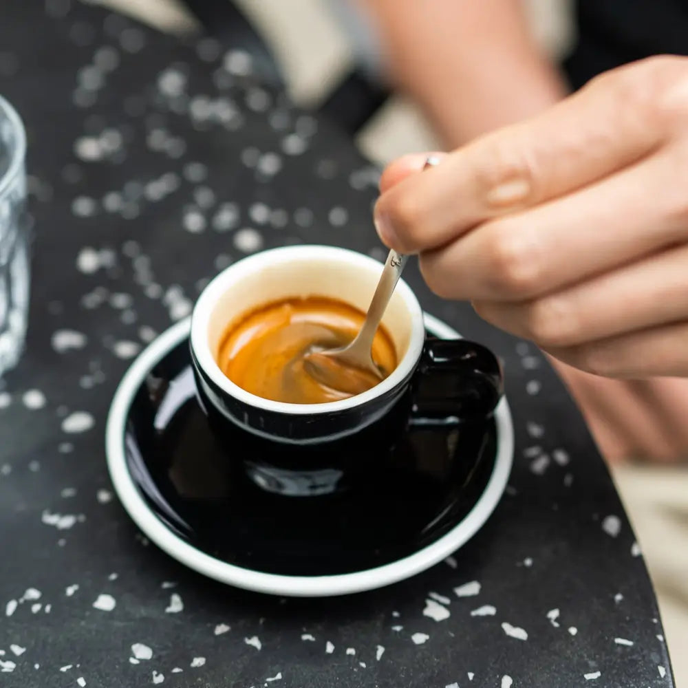 Filiżanka espresso na czarnym spodku, osoba mieszająca kawę łyżeczką przy stoliku z ciemnym, nakrapianym blatem.
