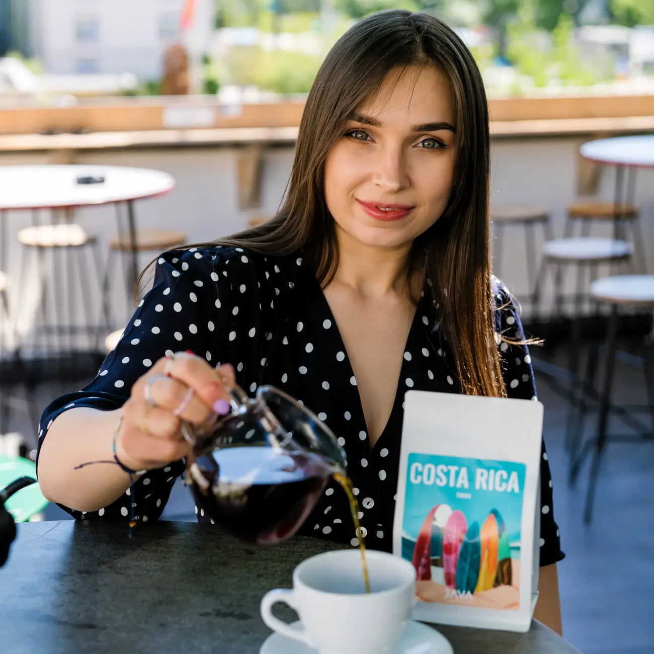 Kawa ziarnista JAVA Costa Rica Tirra 250g – kobieta w kawiarni nalewająca zaparzoną kawę do filiżanki, z opakowaniem kawy na stoliku.