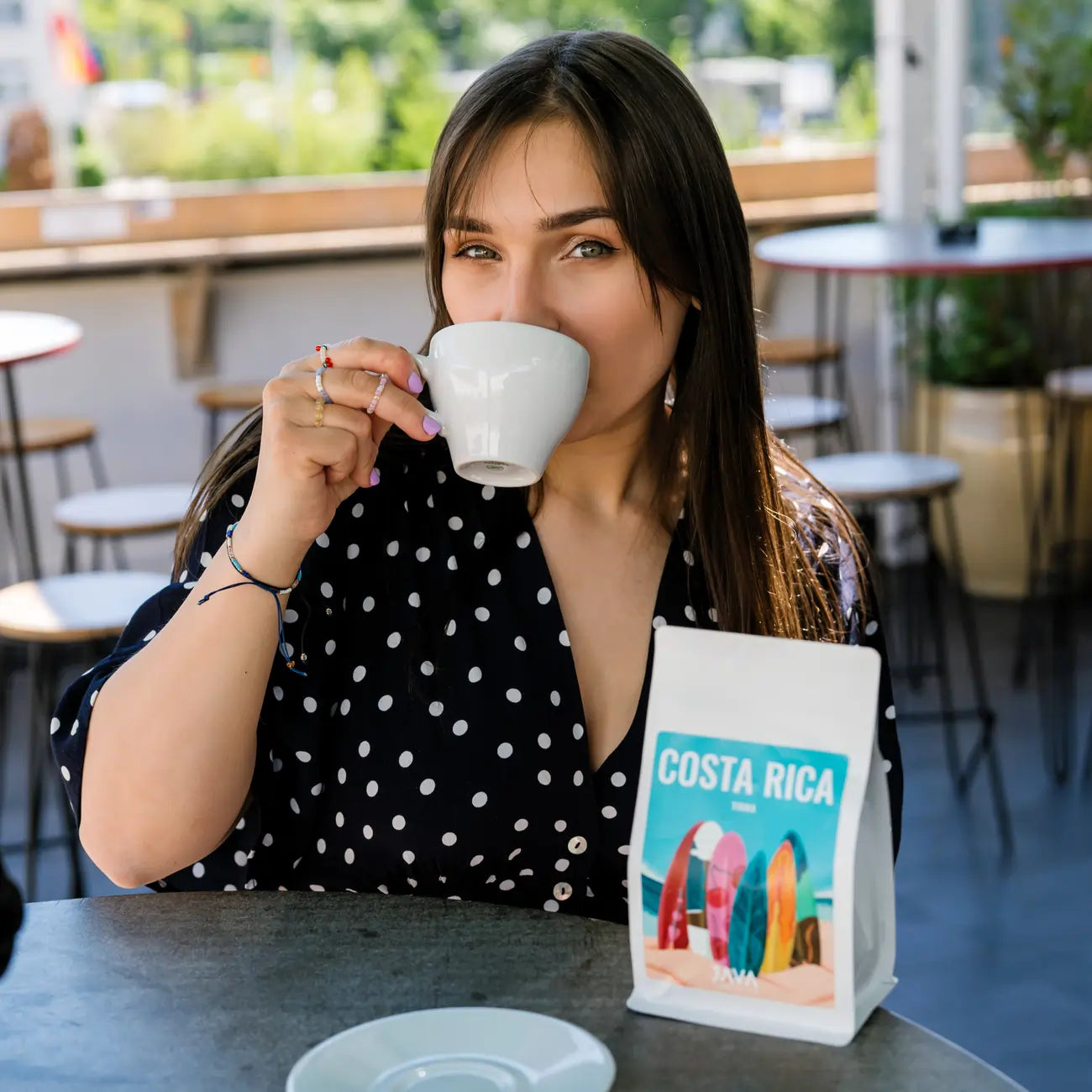 Kawa ziarnista JAVA Costa Rica Tirra 250g – kobieta pijąca kawę w kawiarni, obok opakowanie kawy JAVA na stoliku.