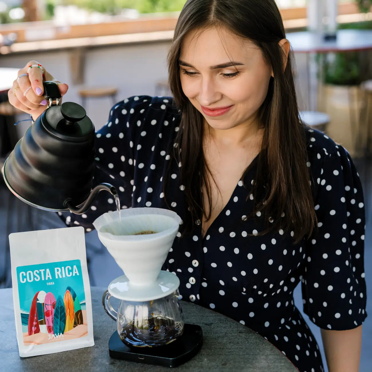 Kawa ziarnista JAVA Costa Rica Tirra 250g – kobieta parzy kawę metodą V60 w kawiarni, obok opakowanie kawy JAVA na stoliku.