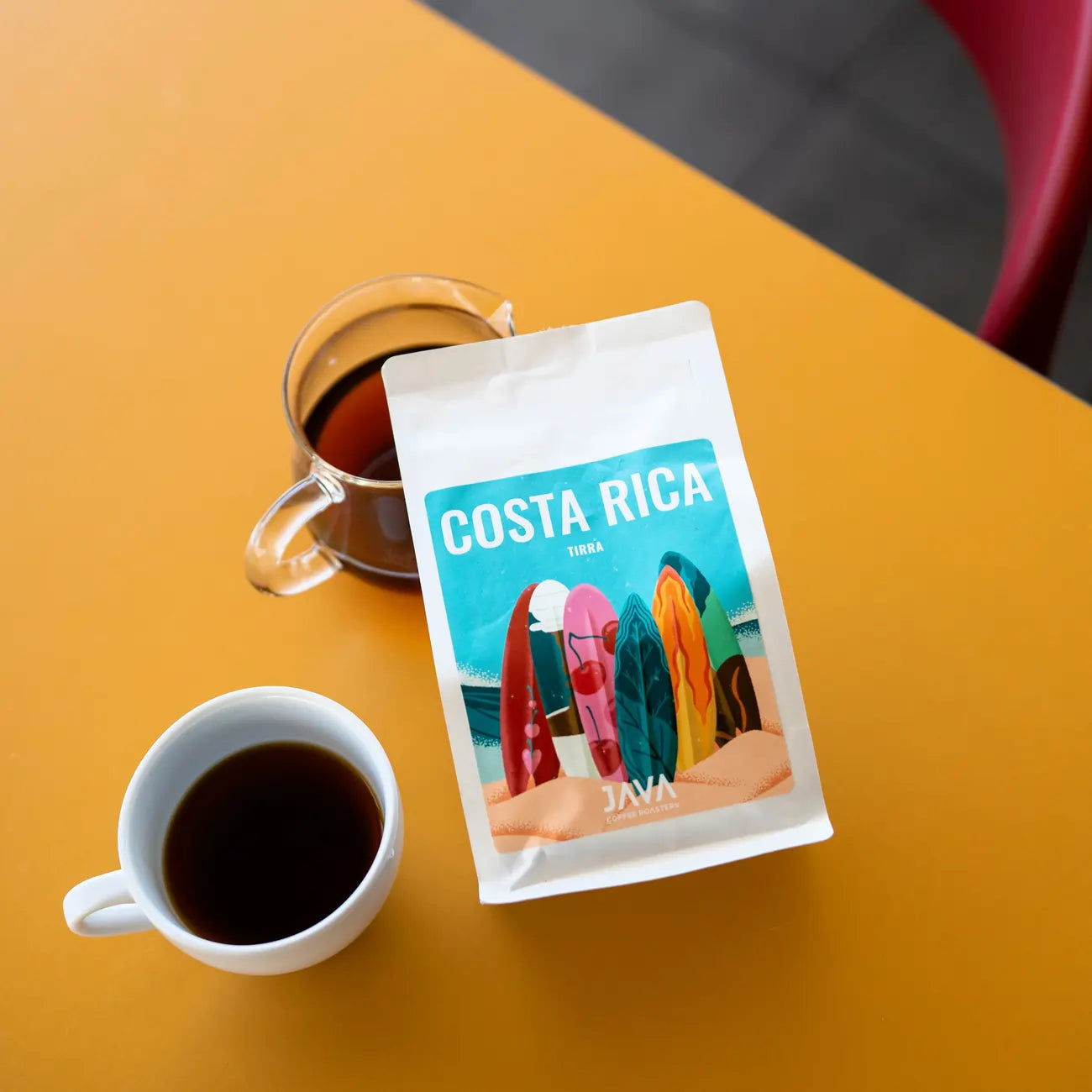Kawa ziarnista JAVA Costa Rica Tirra 250g – opakowanie na stole z zaparzoną kawą w filiżance i dzbanku, ujęcie lifestyle.