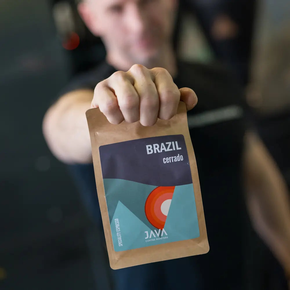 Mężczyzna w koszulce "Good Coffee is a Human Right" prezentuje opakowanie kawy Brazil Cerrado od JAVA Coffee Roasters, opierając je na swoim ramieniu. Palarz kawy w tle palarni. Świeżo palona kawa specialty z Brazylii, idealna do espresso.