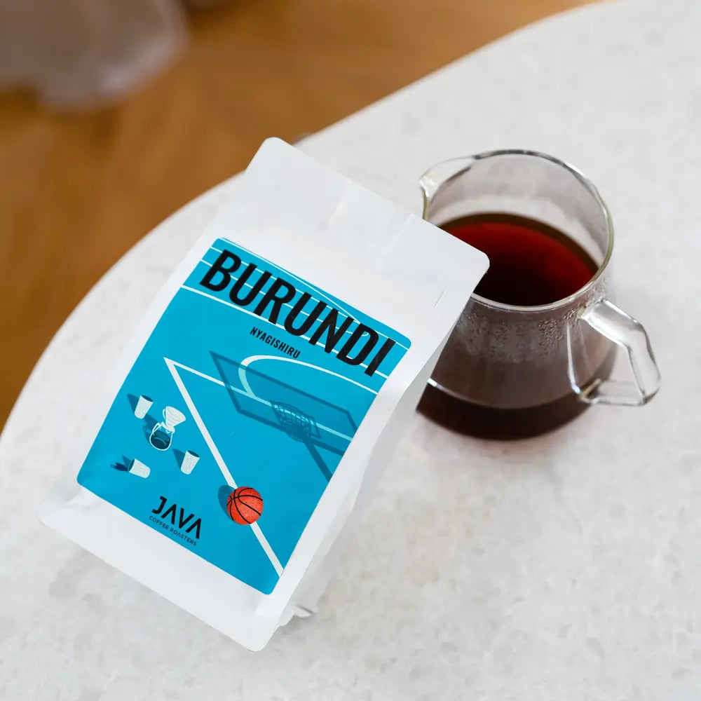 Kawa Burundi Nyagishiru od JAVA Coffee Roasters w białym opakowaniu z niebieską etykietą obok dzbanka z zaparzoną kawą przelewową