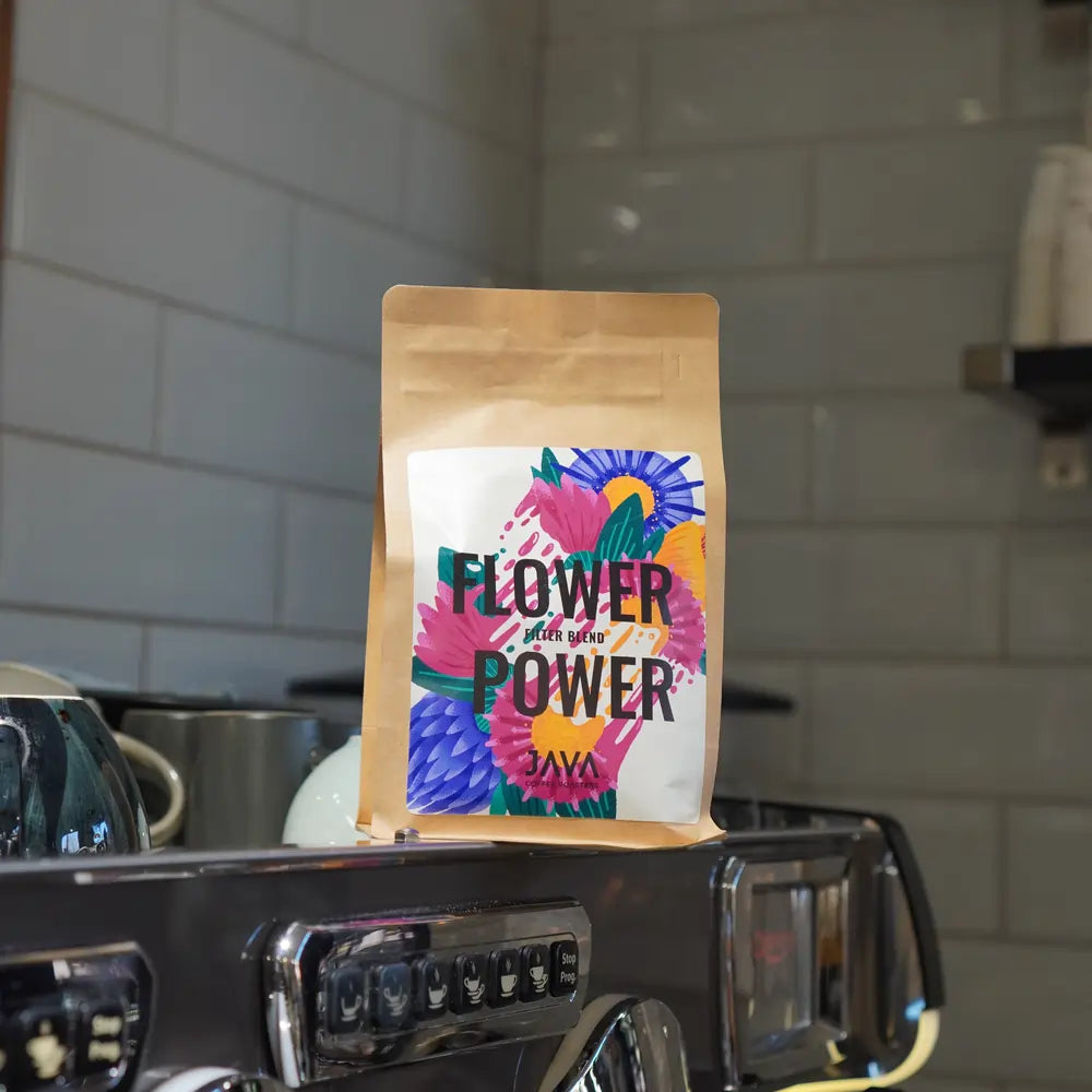 Paczka kawy JAVA Flower Power na ekspresie do kawy w kawiarni, na tle białych kafelków i naczyń na półce.