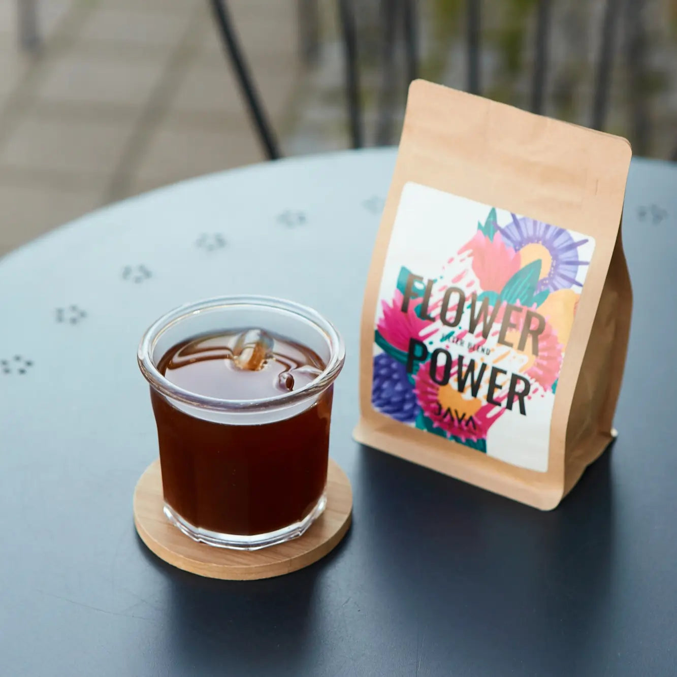 Paczka kawy JAVA Flower Power na metalowym stole obok szklanki z mrożoną kawą na drewnianej podstawce, w tle metalowe krzesła.