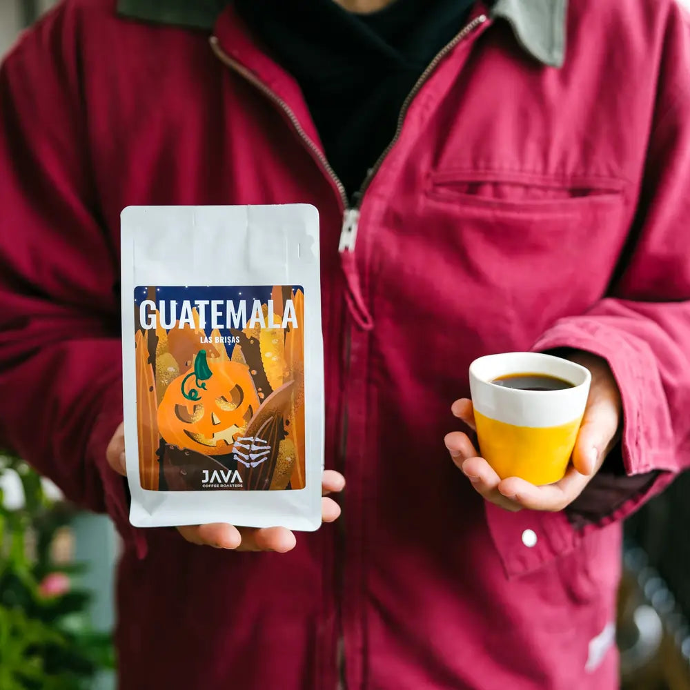 Kawa specialty Guatemala Las Brisas JAVA Coffee Roasters 250g – opakowanie halloweenowe trzymane obok filiżanki kawy