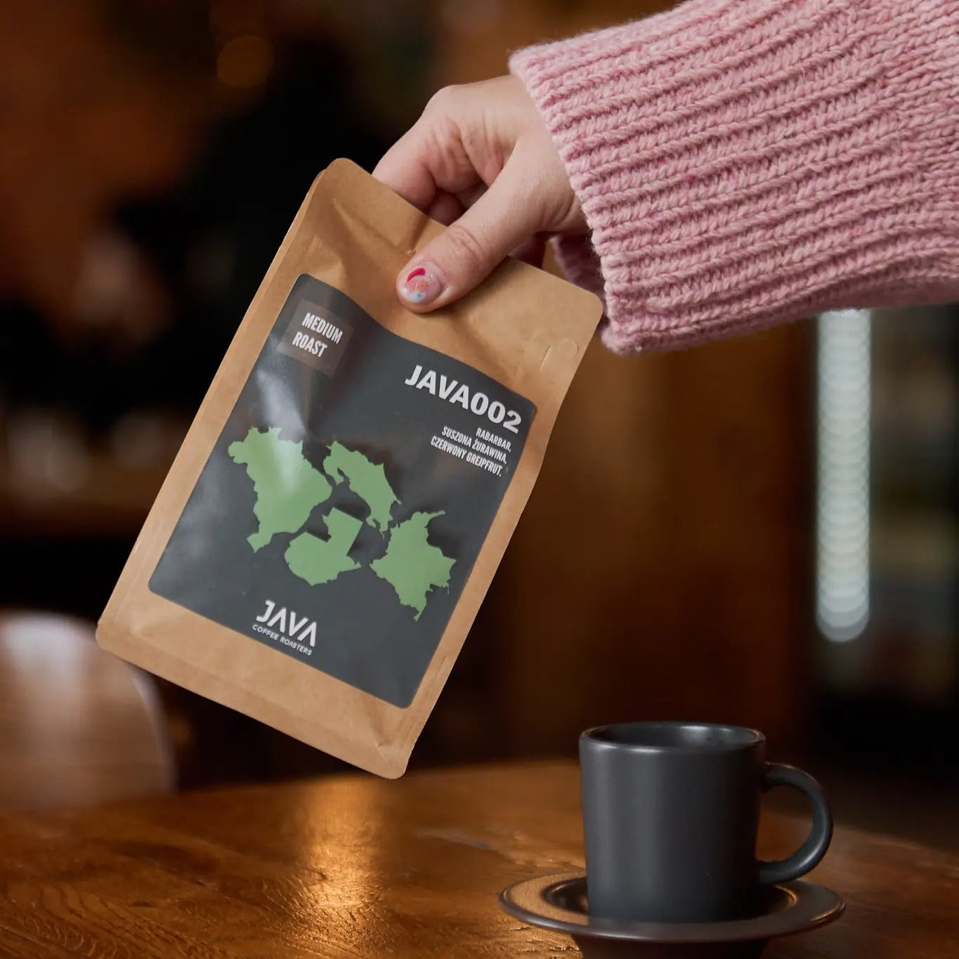 Osoba w różowym swetrze trzyma paczkę kawy JAVA002 Medium Roast nad drewnianym stołem, obok czarnej filiżanki na spodku, z rozmytym tłem kawiarni.