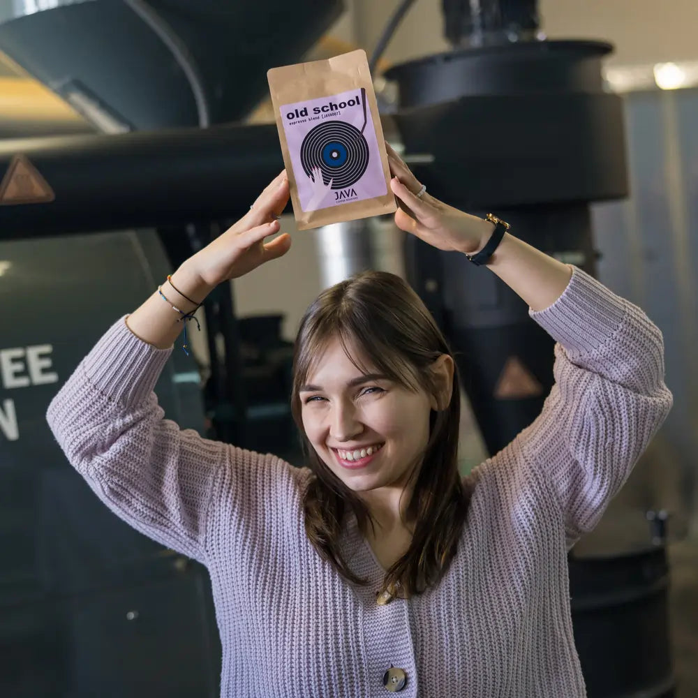 Kobieta w jasnym swetrze trzyma opakowanie kawy Old School Espresso Blend od JAVA Coffee Roasters na tle palarni kawy. Świeżo palona kawa specialty, idealna do klasycznego espresso.