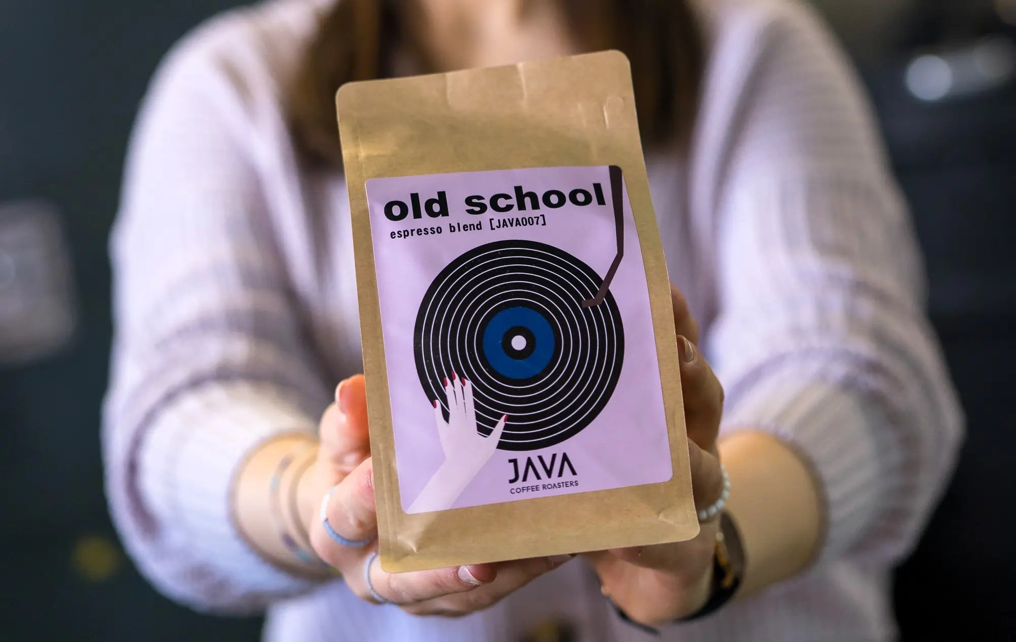 Kobieta w jasnym swetrze trzyma nad głową opakowanie kawy Old School Espresso Blend od JAVA Coffee Roasters, uśmiechając się na tle palarni kawy. Świeżo palona kawa specialty, idealna do espresso.