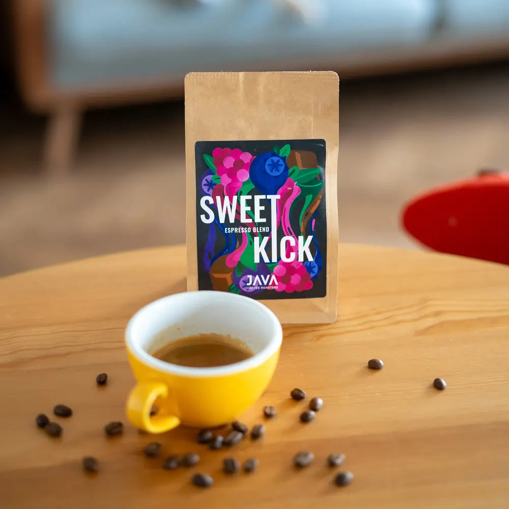 Paczka kawy JAVA Sweet Kick na drewnianym stole, obok żółtej filiżanki z espresso i rozsypanych ziaren kawy.