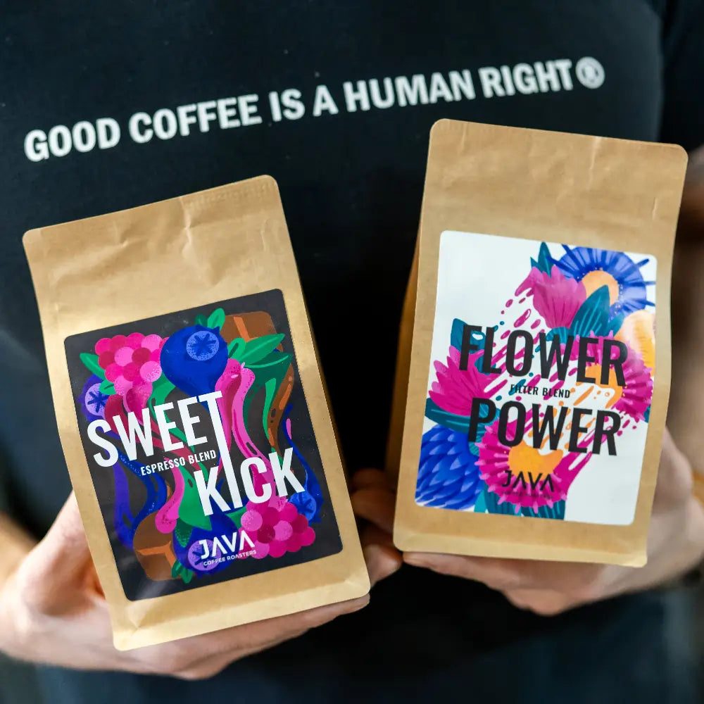 Osoba trzymająca dwie paczki kawy specialty JAVA — Sweet Kick i Flower Power, na tle czarnej koszulki z napisem „GOOD COFFEE IS A HUMAN RIGHT”.