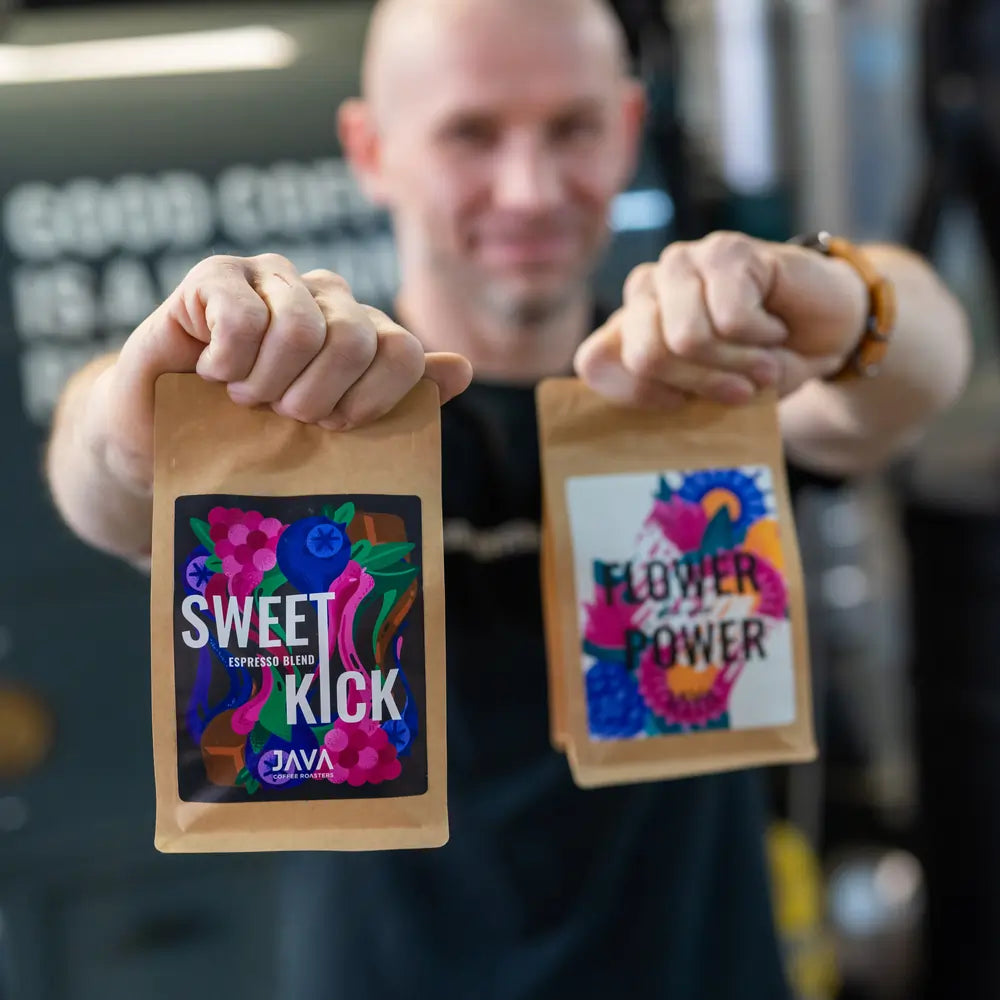 Osoba trzymająca dwie paczki kawy JAVA – Sweet Kick i Flower Power, z kolorowymi etykietami na tle palarni kawy.