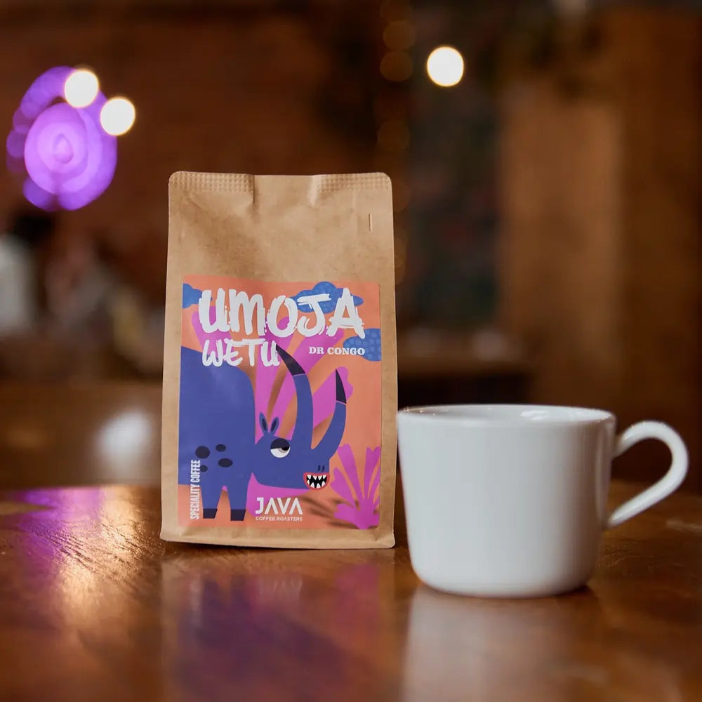Paczka kawy JAVA Umoja Wetu z DR Congo na drewnianym stole, obok białej filiżanki, z rozmytym tłem kawiarni i fioletowym oświetleniem.