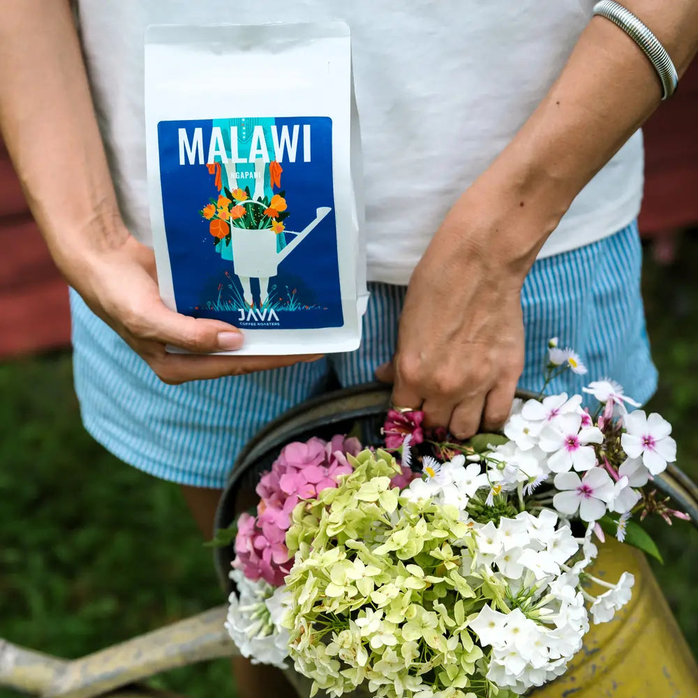 Opakowanie kawy Malawi Ngapani od JAVA Coffee Roasters trzymane w dłoni obok żółtej konewki z kolorowymi kwiatami – oferta kawy specialty dla biur i gastronomii.