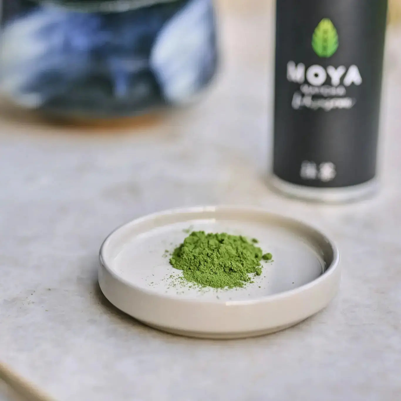 Zbliżenie na sproszkowaną herbatę Moya Matcha Luksusowa na ceramicznym spodku – intensywnie zielony proszek w tle z opakowaniem herbaty.