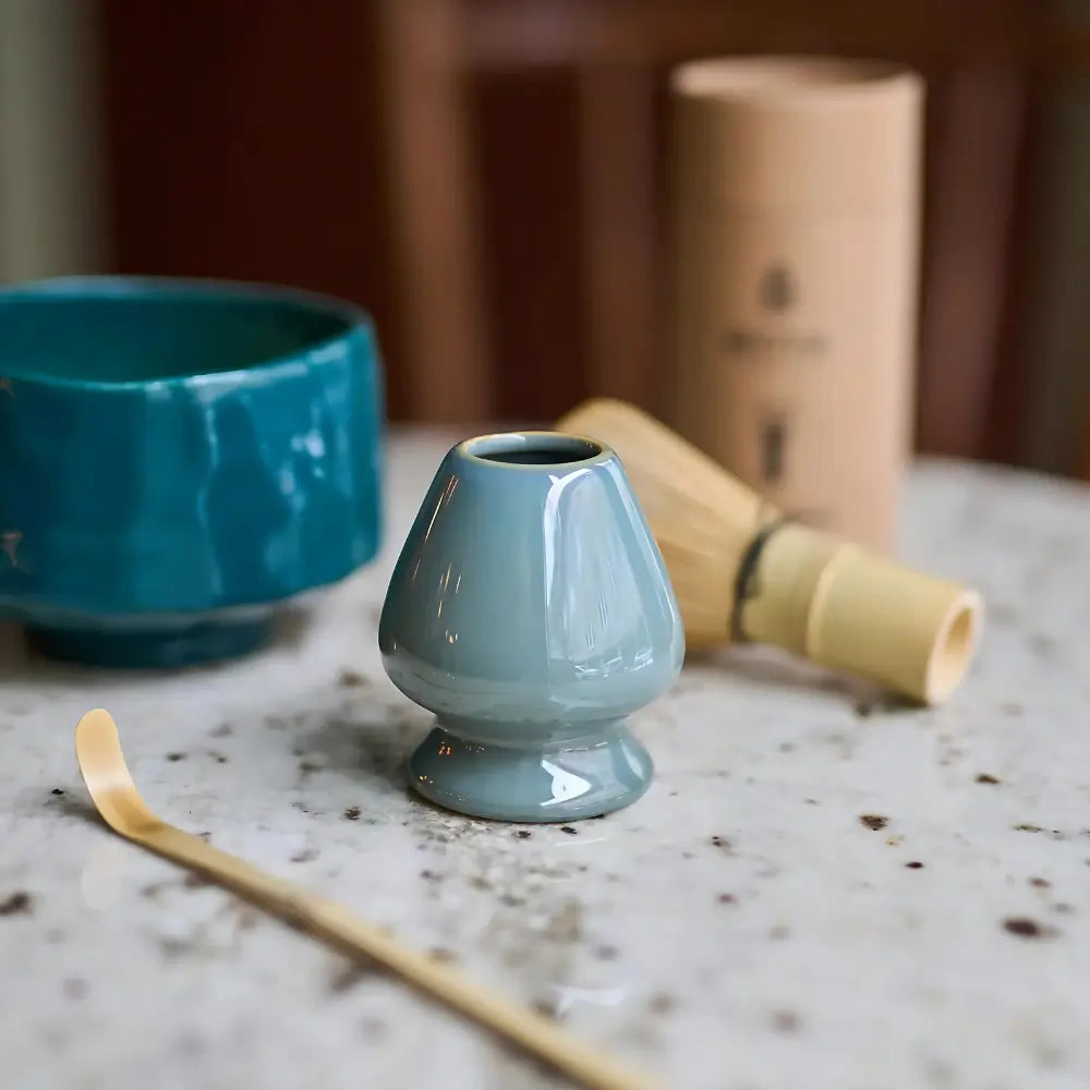 Szaro-zielona podstawka na chasen (naoshi) z porcelany – stylowe akcesorium do matchy na tle miseczki i bambusowej miotełki na marmurowym blacie