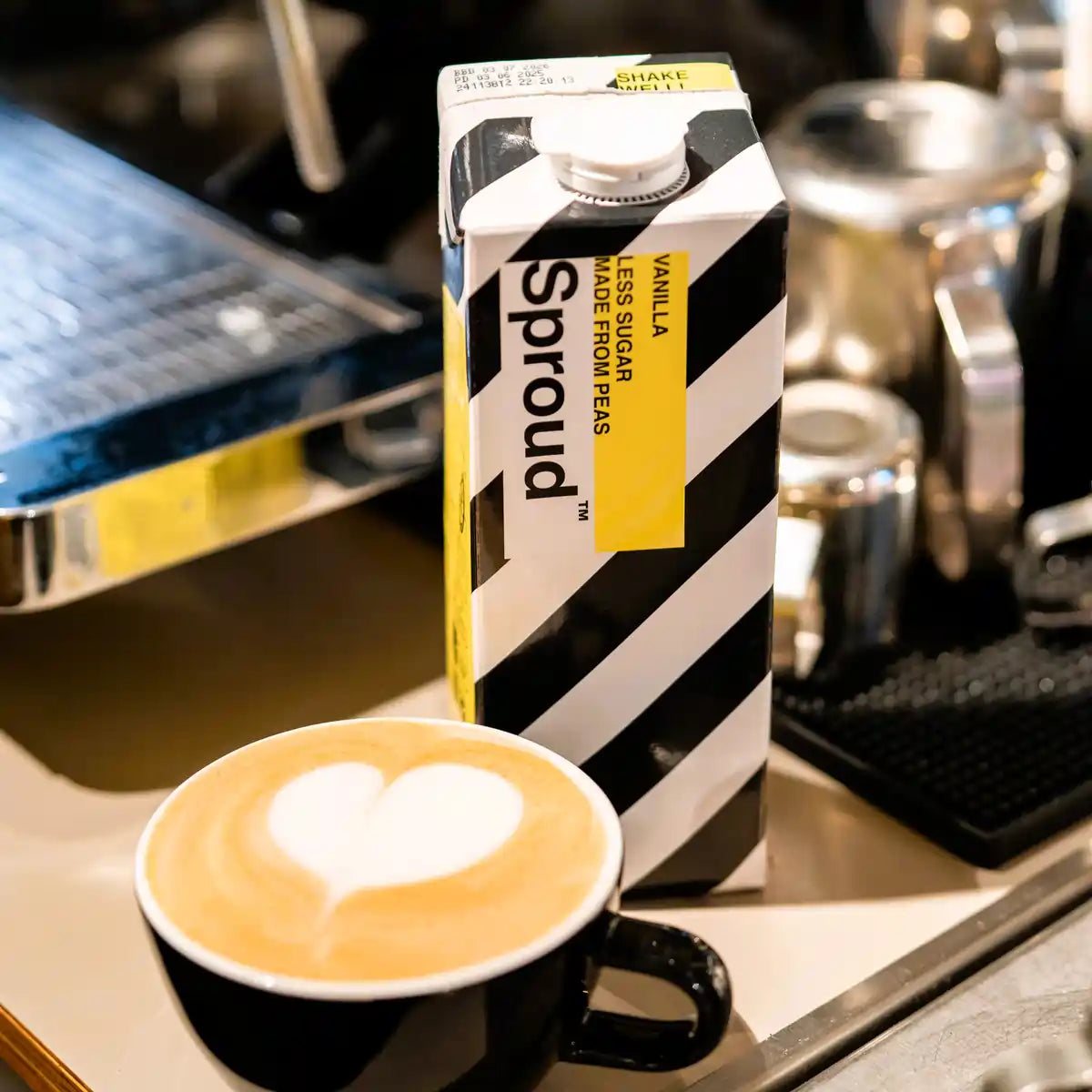 Karton napoju roślinnego Sproud Vanilla 1L obok filiżanki kawy latte z latte art – wegański napój z grochu o smaku waniliowym, idealny do kaw mlecznych.