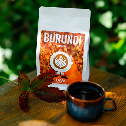 Opakowanie kawy specialty Burundi Burunga od JAVA Coffee Roasters ustawione na drewnianym stole obok filiżanki czarnej kawy, w otoczeniu naturalnej zieleni.