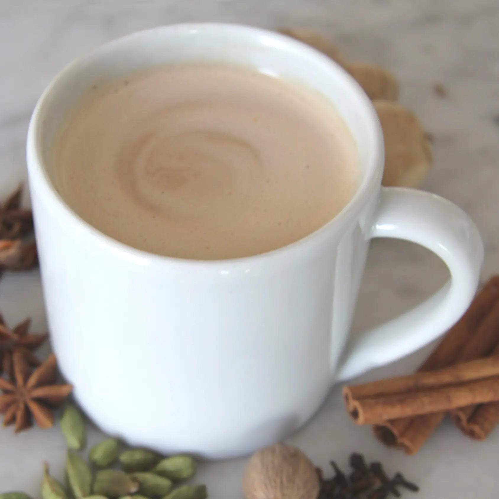 Kubek chai latte w białej filiżance otoczony przyprawami – cynamon, anyż, kardamon, imbir