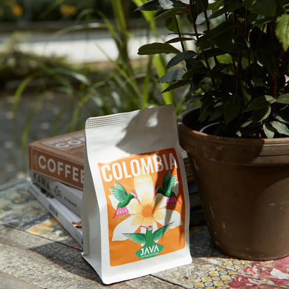 Kawa Colombia Tolima JAVA Coffee Roasters – opakowanie ustawione obok książek o kawie i doniczki z rośliną na rustykalnym stole