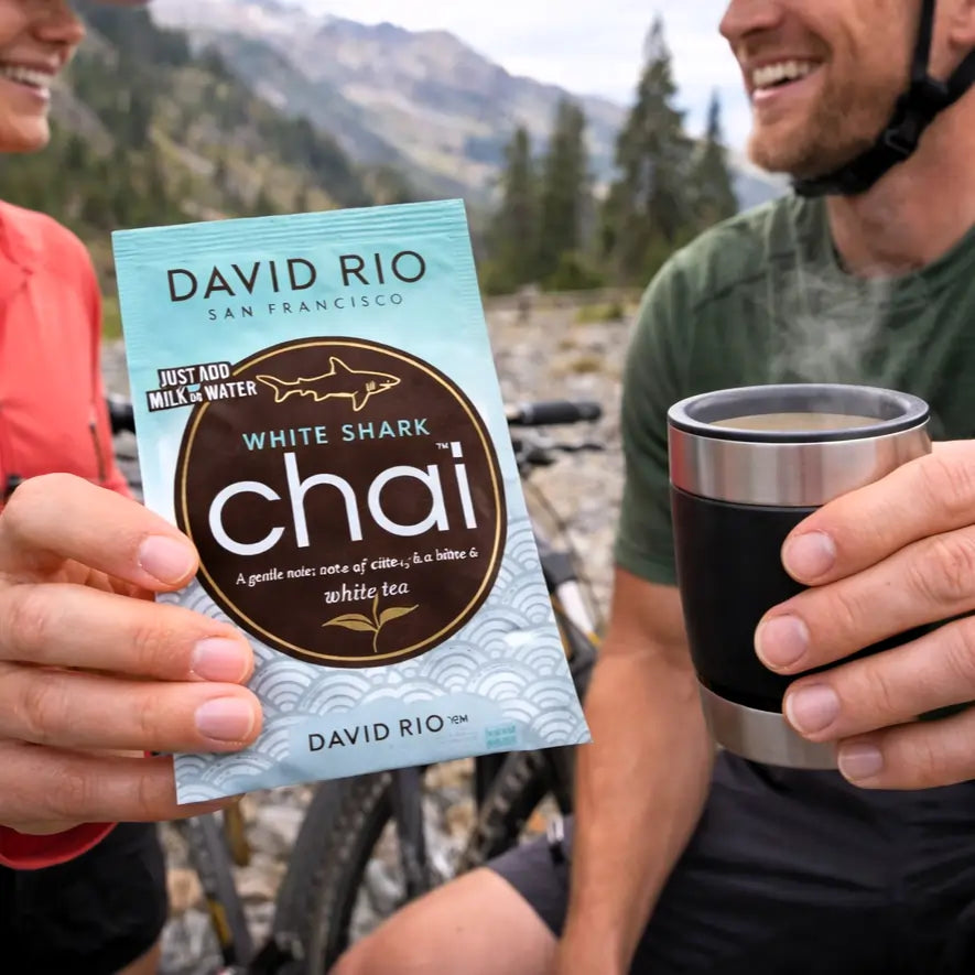 White Shark Chai David Rio – delikatne chai na bazie białej herbaty w saszetce, trzymane w dłoni podczas górskiej przerwy, obok kubka gorącego napoju, lifestyle outdoor