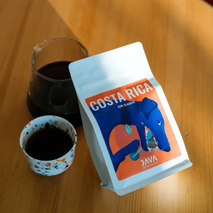 Kawa specialty Costa Rica Don Claudio od JAVA Coffee Roasters – opakowanie z filiżanką czarnej kawy i karafką na drewnianym stole, zdjęcie lifestyle
