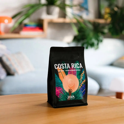 Kawa specialty JAVA Coffee Costa Rica Las Lajas Black Diamond Natural w czarnym opakowaniu ustawiona na drewnianym stole, w tle jasne wnętrze z roślinami.