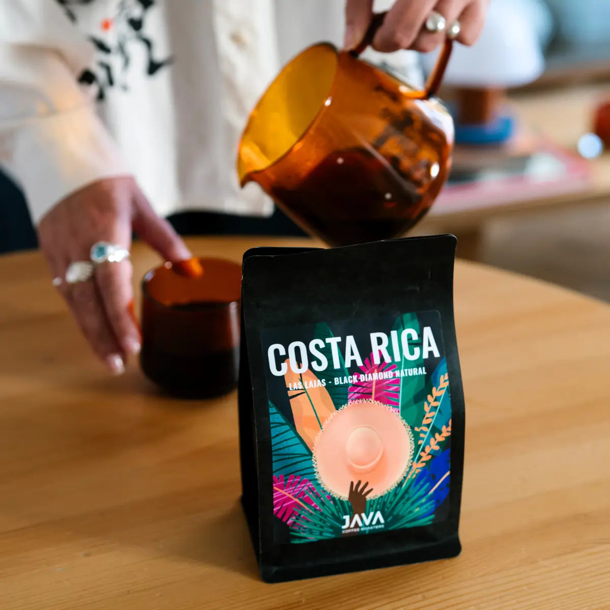Kawa specialty JAVA Coffee Costa Rica Las Lajas Black Diamond Natural w czarnym opakowaniu na stole, w tle osoba nalewająca kawę z bursztynowego dzbanka do filiżanki