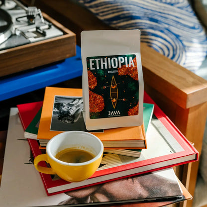 Kawa JAVA Coffee Roasters Ethiopia Chelektu stojąca na stosie książek obok filiżanki espresso w żółtej filiżance w przytulnym, domowym wnętrzu.