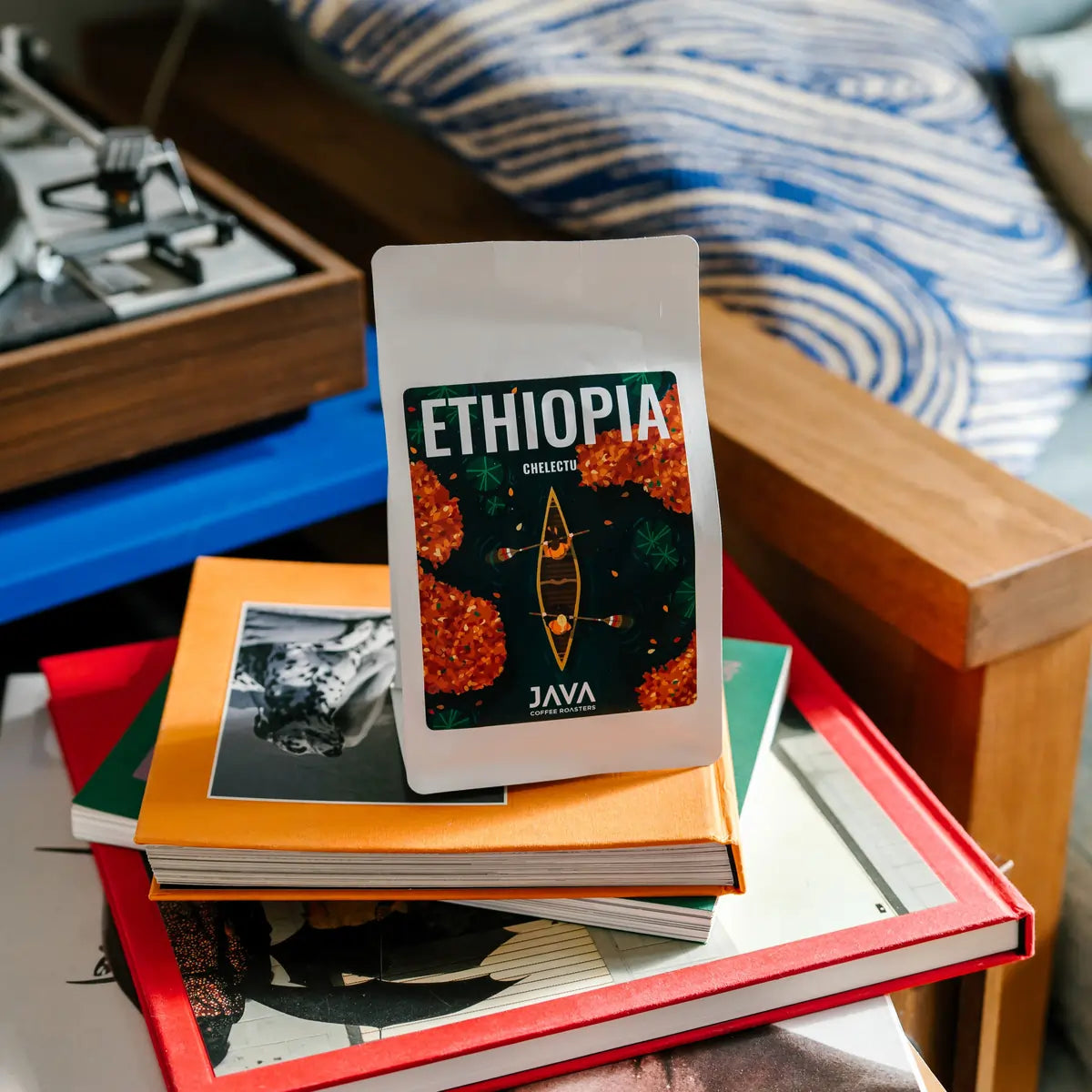 Opakowanie kawy JAVA Coffee Roasters Ethiopia Chelektu stojące na stosie kolorowych książek w domowym wnętrzu z gramofonem w tle.