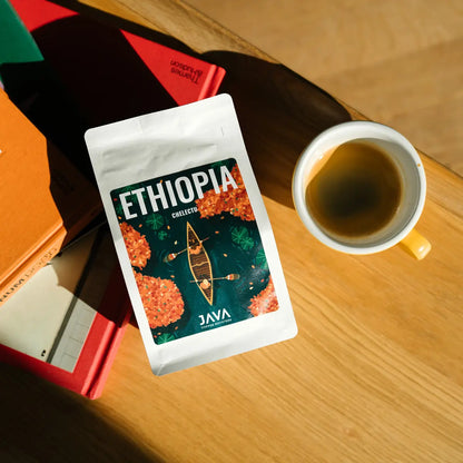 Paczka kawy JAVA Coffee Roasters Ethiopia Chelektu leżąca na stole obok filiżanki kawy i książek w ciepłym świetle poranka.