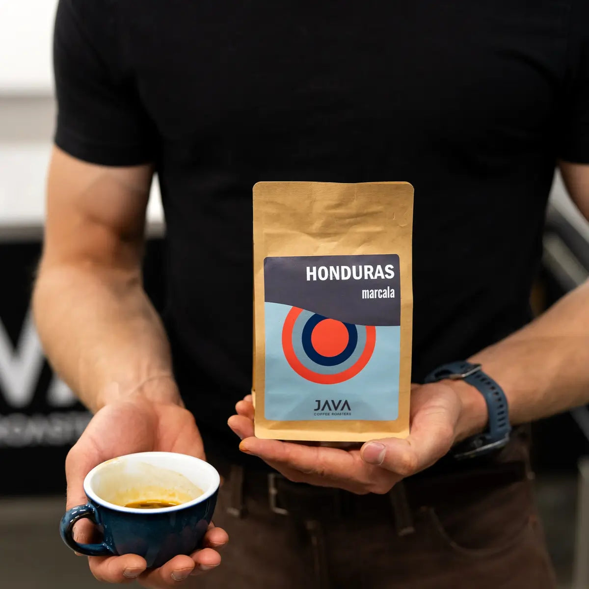 Trener kawowy trzymający opakowanie kawy specialty Honduras Marcala od JAVA Coffee Roasters oraz filiżankę świeżo zaparzonego espresso.