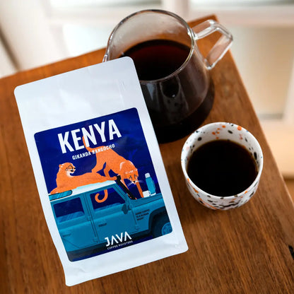 Kawa ziarnista Kenya Gikanda Kangoch od JAVA Coffee Roasters – opakowanie kawy na drewnianym stole, w tle szklany dzbanek z zaparzoną kawą oraz filiżanka czarnej kawy, naturalne światło.