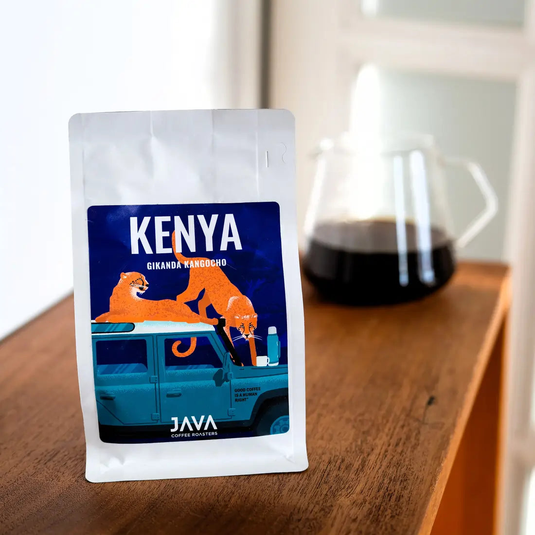 Kawa ziarnista Kenya Gikanda Kangoch od JAVA Coffee Roasters – opakowanie kawy na drewnianym stole, w tle szklany dzbanek z zaparzoną czarną kawą, jasne naturalne światło.