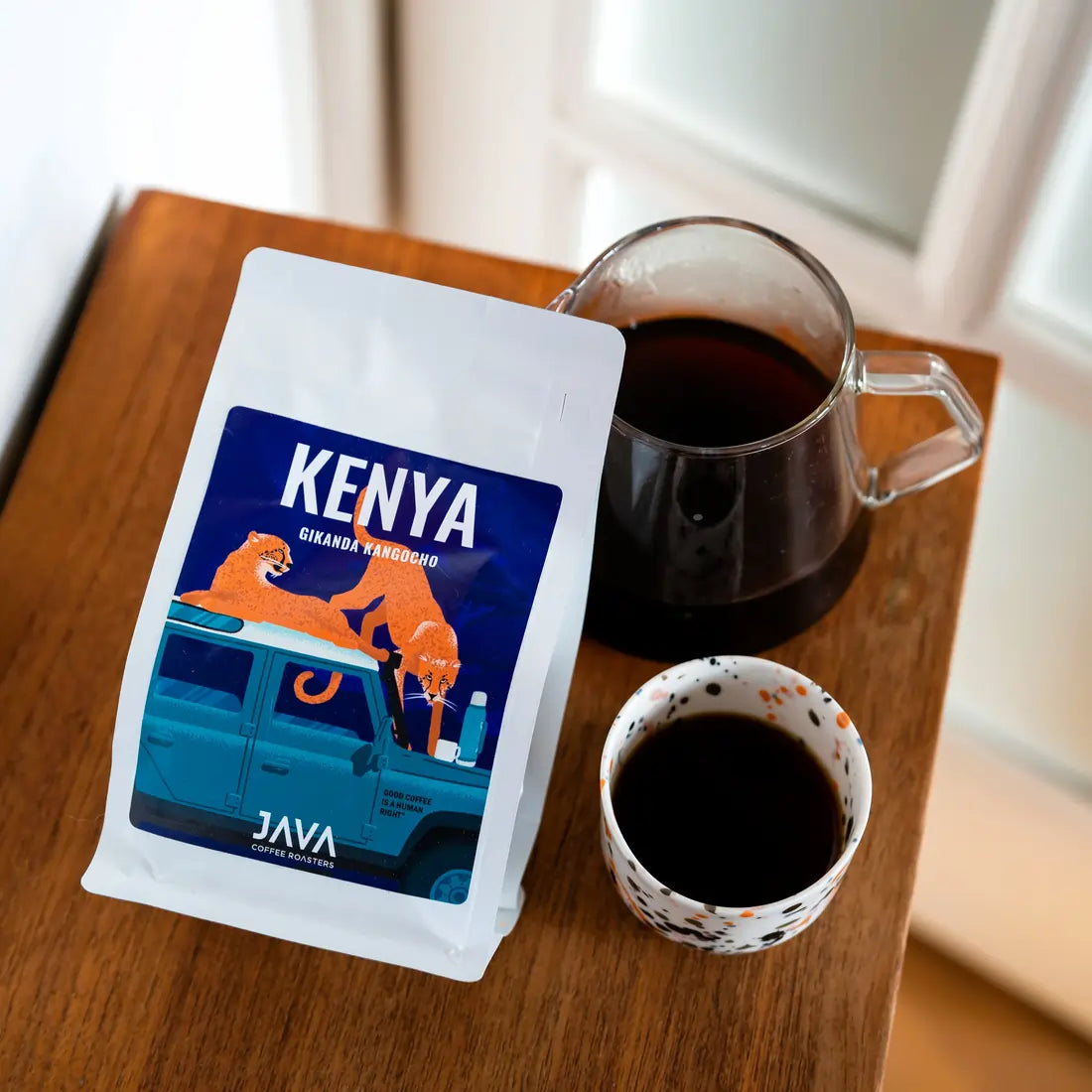 Kawa ziarnista Kenya Gikanda Kangoch od JAVA Coffee Roasters – opakowanie kawy, szklany dzbanek z zaparzoną czarną kawą oraz filiżanka kawy na drewnianym stole, ujęcie z góry.