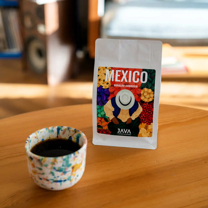 Kawa specialty Mexico Donaldo Ambrosio od JAVA Coffee Roasters – opakowanie kawy do metod przelewowych obok ceramicznego kubka z czarną kawą, zdjęcie lifestyle w domowym wnętrzu.
