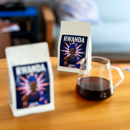 Dwa opakowania kawy specialty Rwanda Cocatu od JAVA Coffee Roasters oraz szklany serwer z zaparzoną kawą. Zdjęcie prezentuje opakowanie produktu i sposób podania kawy metodą przelewową w naturalnym, domowym otoczeniu.