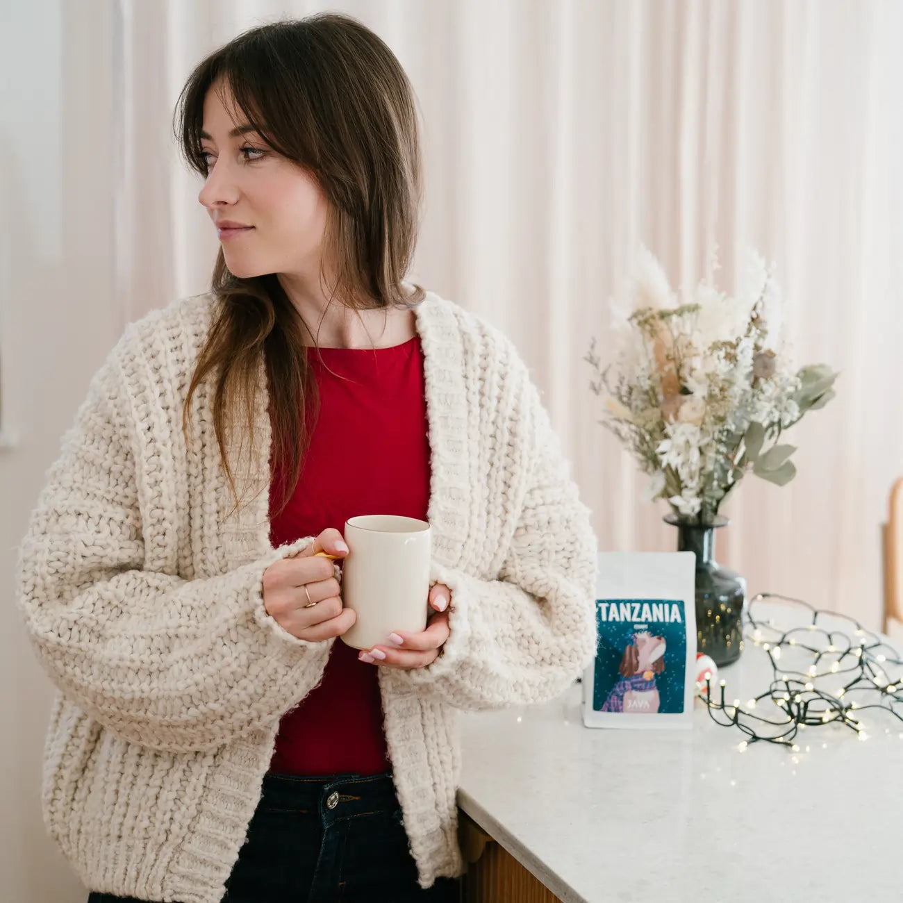 Kobieta trzyma kubek z kawą w jasnym, przytulnym wnętrzu; w tle widoczne opakowanie JAVA Coffee Tanzania Itumpi.