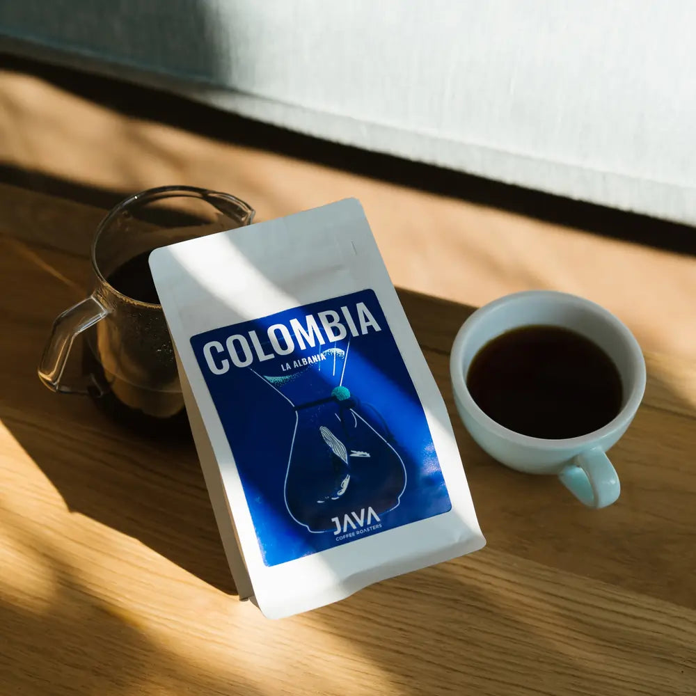 Kawa speciality Java Coffee Roasters Colombia La Albania w białym opakowaniu obok filiżanki i dzbanka z kawą w naturalnym świetle