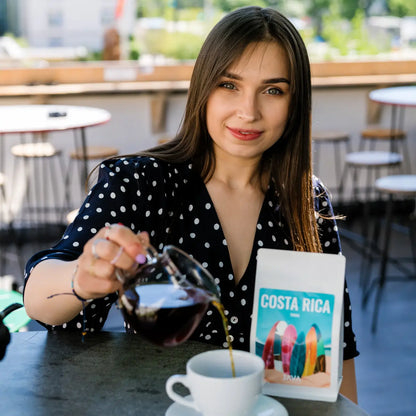 Kawa ziarnista JAVA Costa Rica Tirra 250g – kobieta w kawiarni nalewająca zaparzoną kawę do filiżanki, z opakowaniem kawy na stoliku.