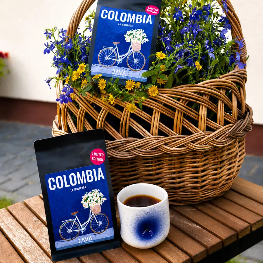 Kawa ziarnista Colombia La Molienda JAVA Coffee w wiklinowym koszu z kwiatami obok filiżanki świeżo zaparzonej kawy.