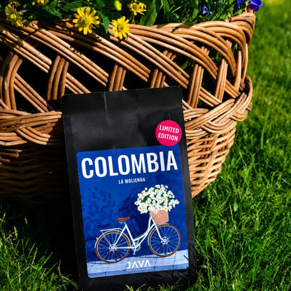 Opakowanie kawy ziarnistej Colombia La Molienda JAVA Coffee ustawione na trawie przed wiklinowym koszem z kwiatami.