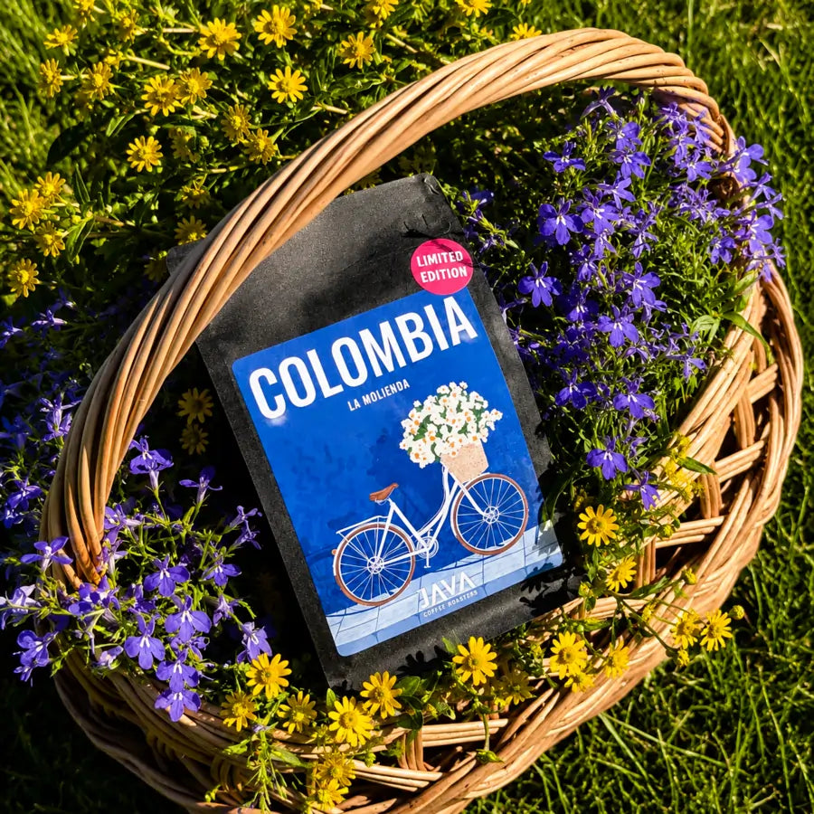 Opakowanie kawy ziarnistej Colombia La Molienda JAVA Coffee w wiklinowym koszu wypełnionym fioletowymi i żółtymi kwiatami.