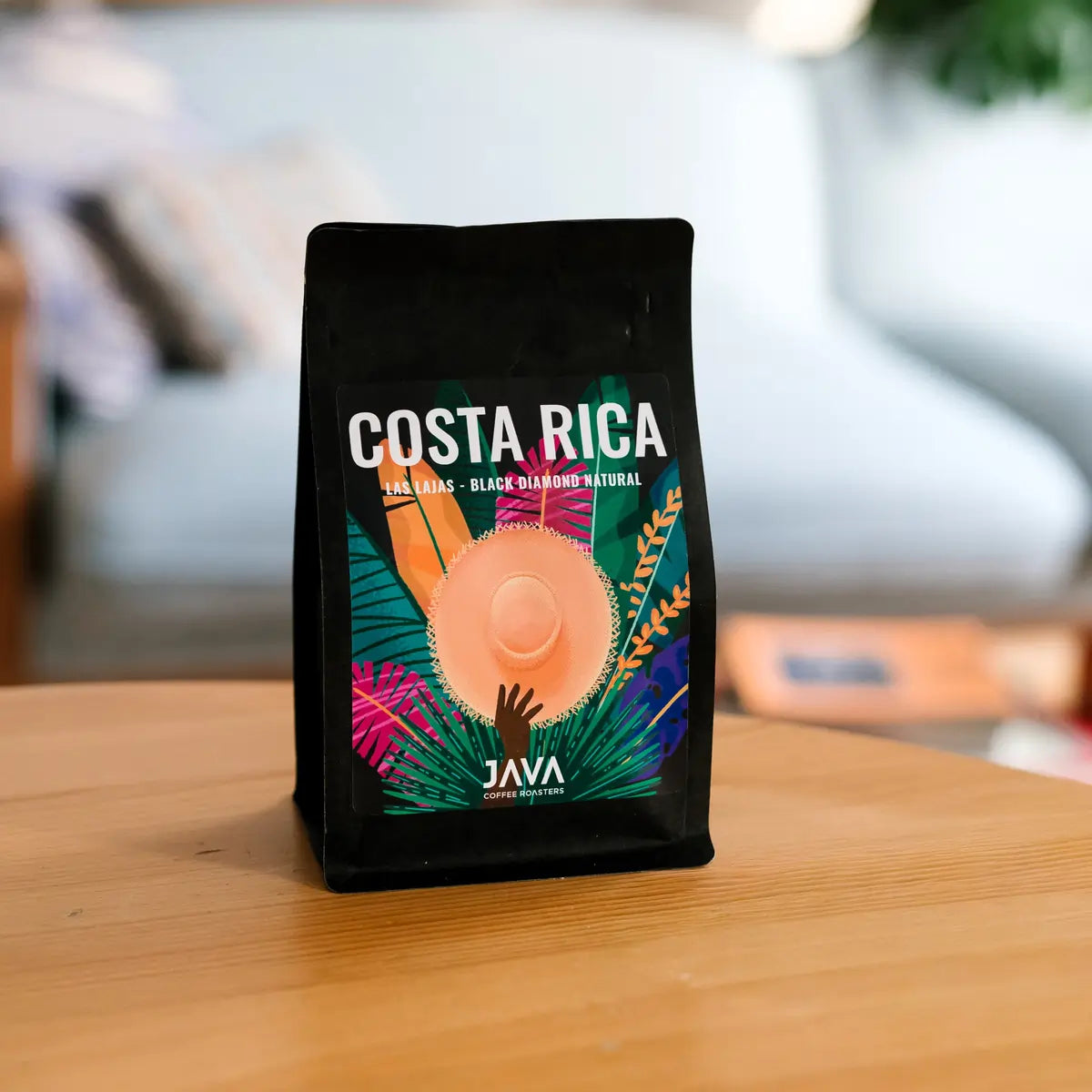 Kawa specialty JAVA Coffee Costa Rica Las Lajas Black Diamond Natural w czarnym opakowaniu stojąca na drewnianym stole, w tle jasne wnętrze kawiarni